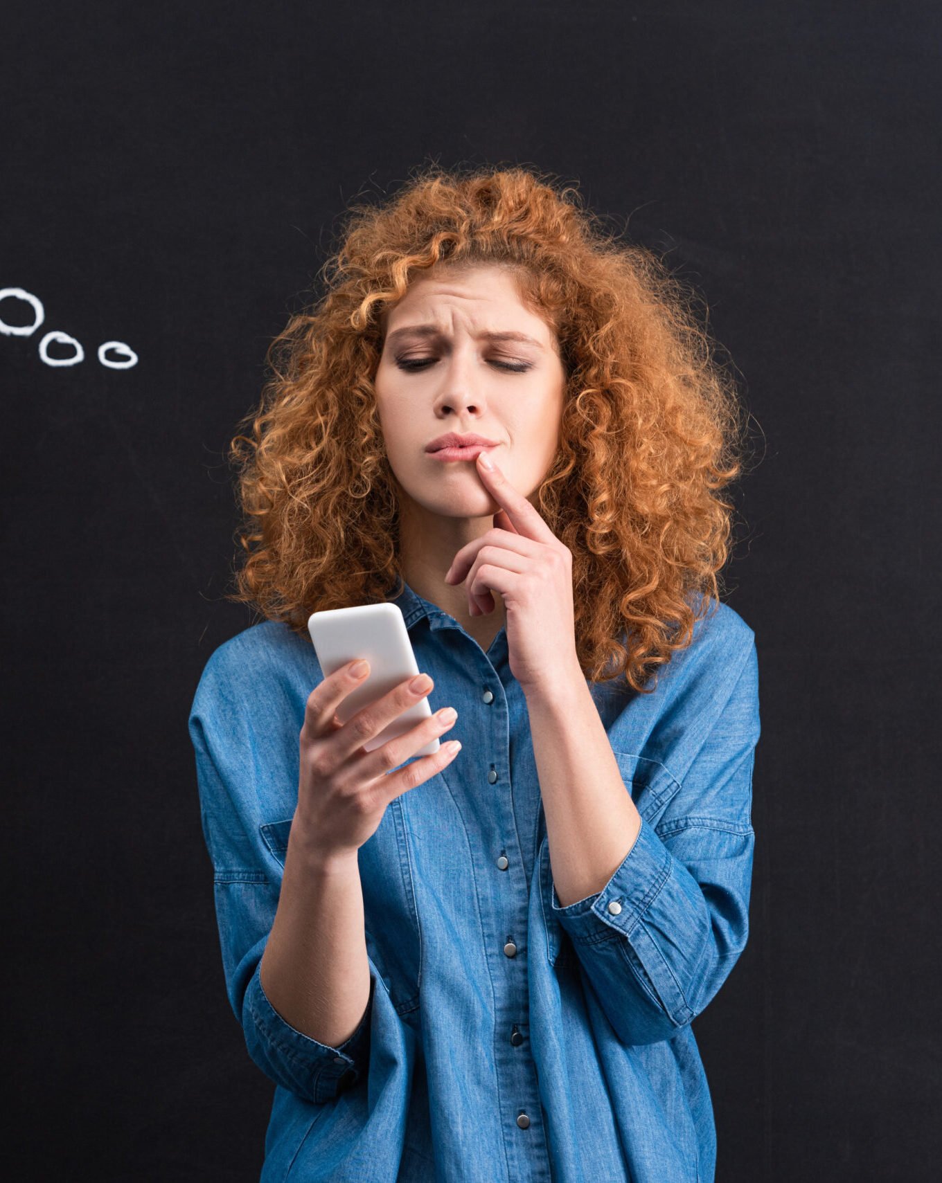 pensive woman using smartphone, question marks in thought bubble on blackboard behind